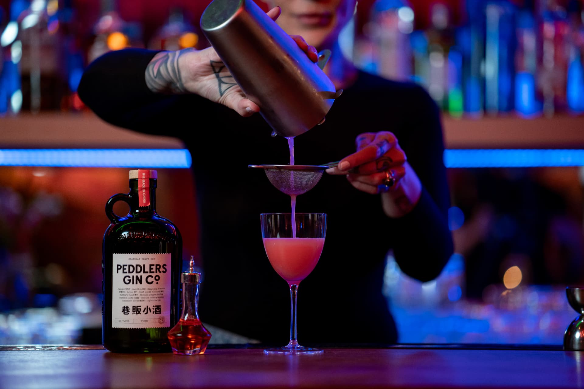 Bartender double-straining a cocktail into a stemmed glass at a back bar